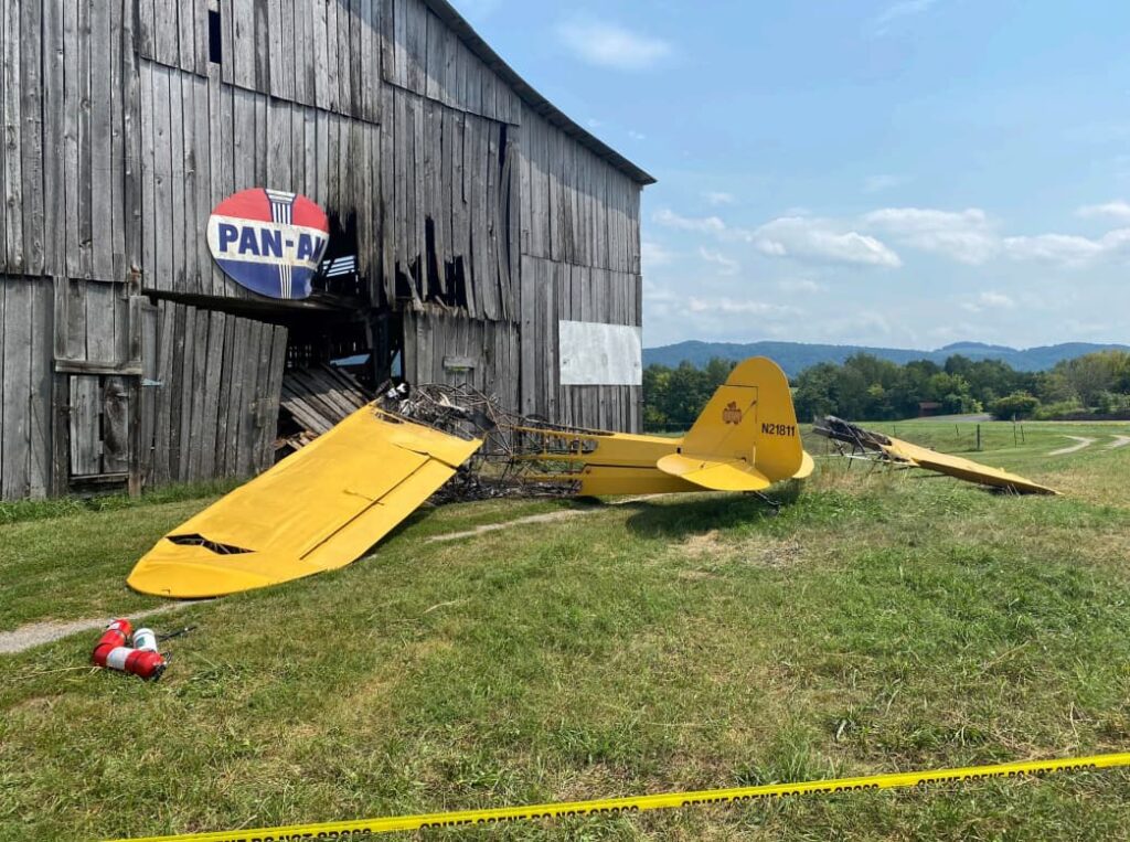 Wide-angle view of the crash site showing the yellow Piper J3 Cub (N21811) wreckage in front of a wooden barn with a Pan-Am sign. The aircraft is partially burned and surrounded by caution tape and fire extinguishers on a grassy field.